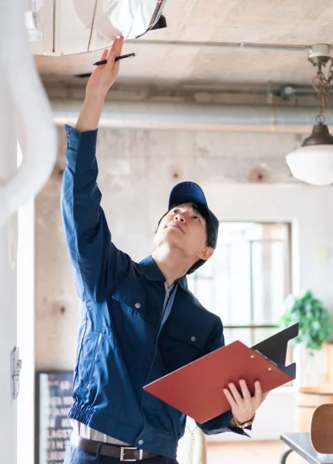 Technician in a blue uniform inspecting a ceiling air conditioning unit while holding a clipboard.