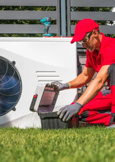 HVAC technician in red uniform servicing a heat pump unit outdoors.